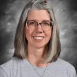 Una mujer con gafas y el pelo gris hasta los hombros sonríe, vestida con una camiseta gris y un collar, con un fondo nublado.